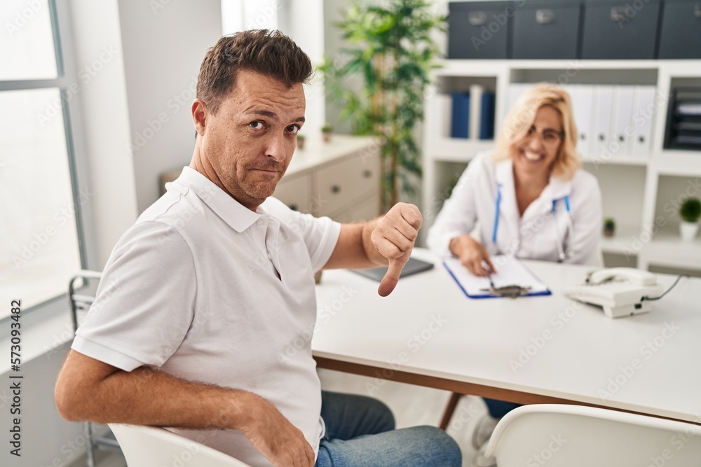 Hispanic man at the doctor with angry face, negative sign showing dislike with thumbs down, rejection concept