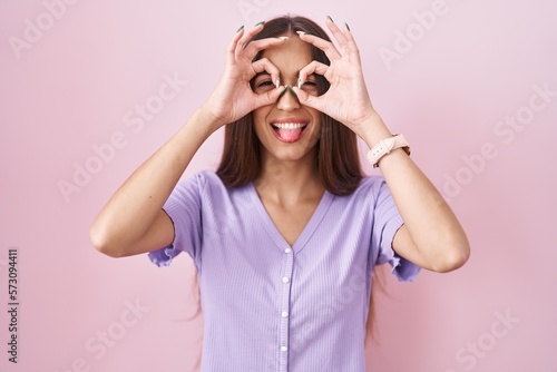 Young hispanic woman with long hair standing over pink background doing ok gesture like binoculars sticking tongue out, eyes looking through fingers. crazy expression. © Krakenimages.com
