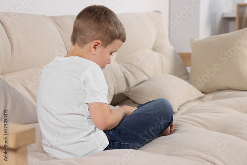 Boy with poor posture sitting on beige sofa in room. Symptom of scoliosis