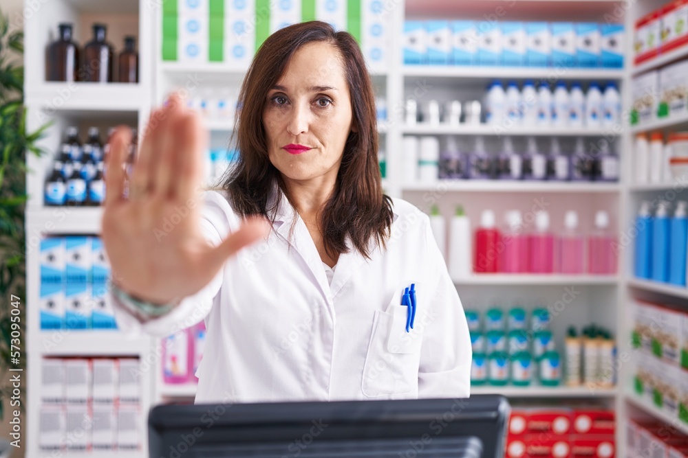 Middle age brunette woman working at pharmacy drugstore doing stop sing with palm of the hand. warning expression with negative and serious gesture on the face.