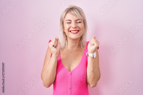 Young caucasian woman standing over pink background excited for success with arms raised and eyes closed celebrating victory smiling. winner concept.