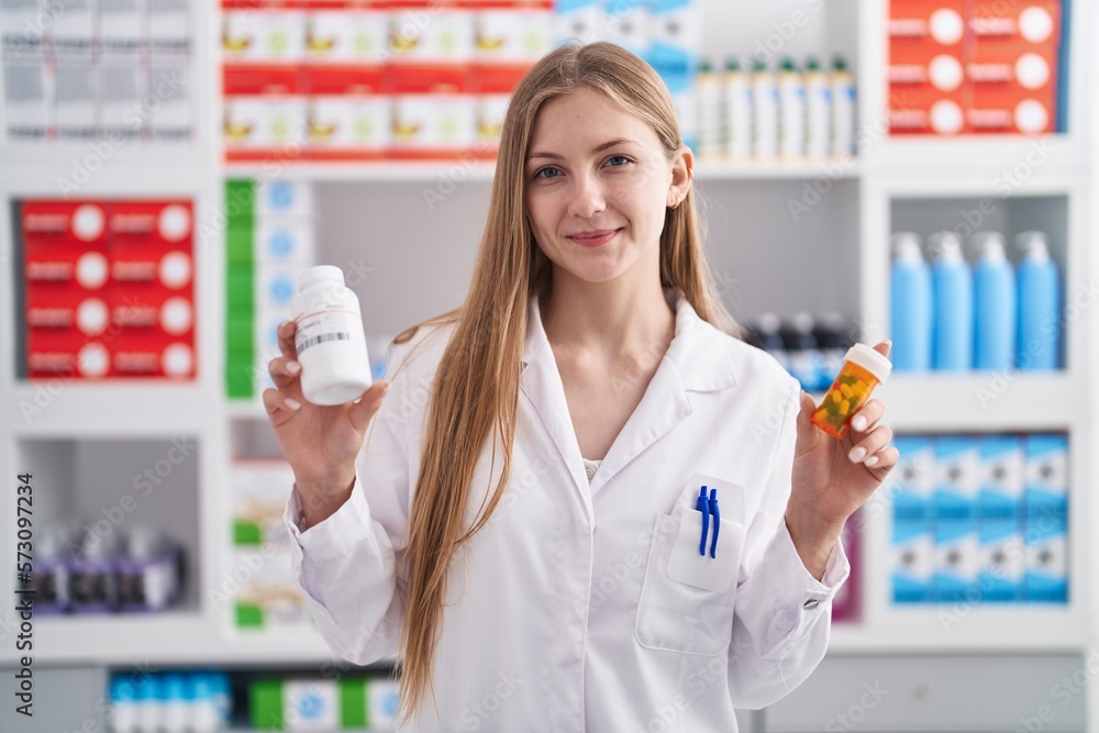 Young caucasian woman pharmacist smiling confident holding pills bottles at pharmacy