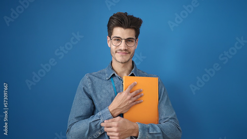 Young hispanic man smiling confident holding book over isolated blue background