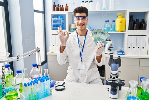Young hispanic man working at scientist laboratory holding brazilian reals doing ok sign with fingers  smiling friendly gesturing excellent symbol