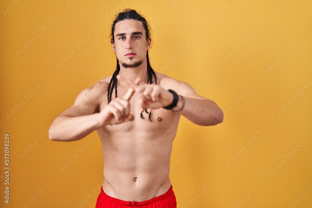 Hispanic man with long hair standing shirtless over yellow background rejection expression crossing fingers doing negative sign