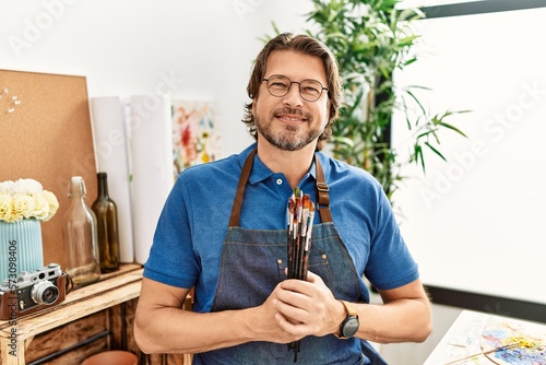 Middle age caucasian man smiling confident holding paintbrushes at art studio