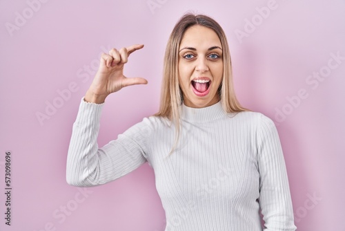 Young blonde woman wearing white sweater over pink background smiling and confident gesturing with hand doing small size sign with fingers looking and the camera. measure concept.