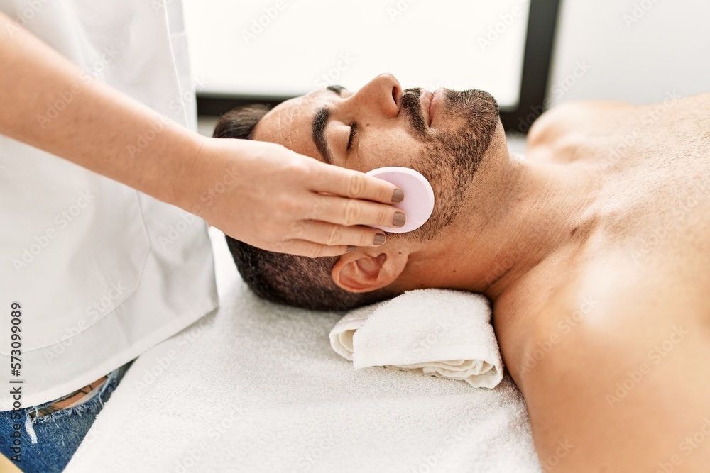 Young hispanic man having clean facial treatment using sponge at beauty center
