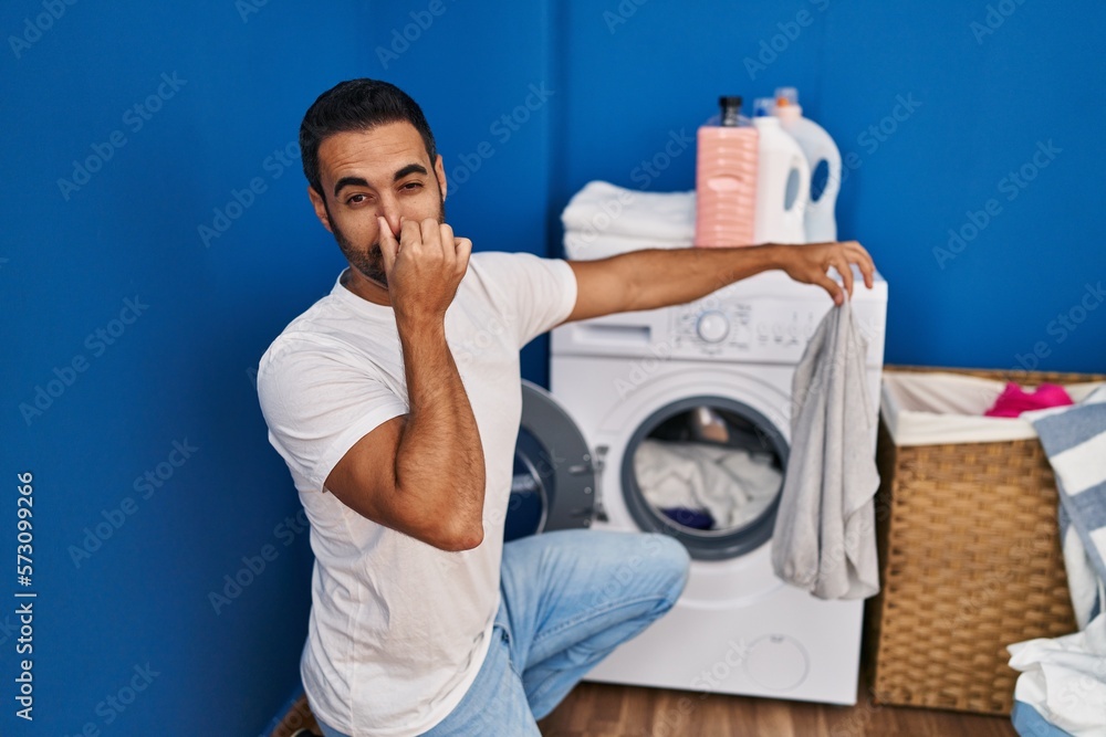 Obraz premium Young hispanic man holding dirty clothe closing nose at laundry room