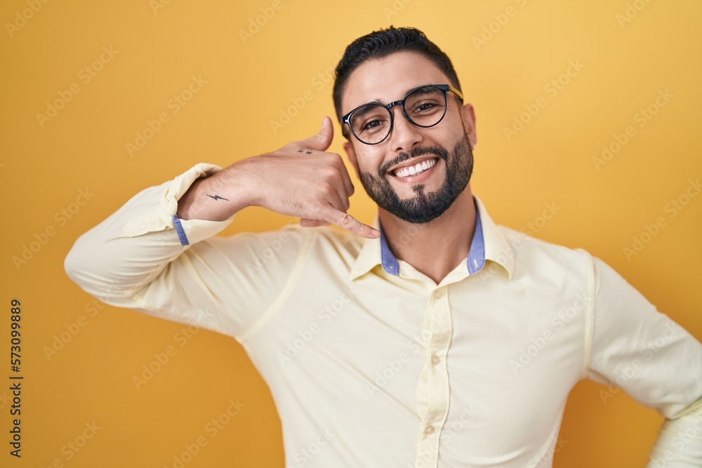 Hispanic young man wearing business clothes and glasses smiling doing phone gesture with hand and fingers like talking on the telephone. communicating concepts. Hispanic young man wearing business clothes and glasses smiling doing phone gesture with hand and fingers like talking on the telephone. communicating concepts.
