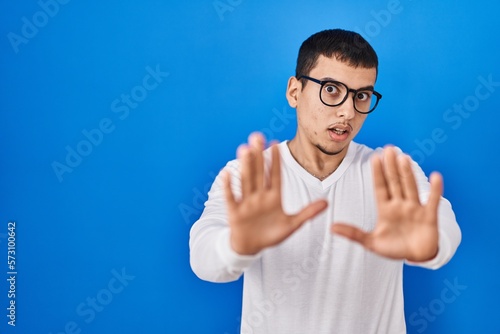 Young arab man wearing casual white shirt and glasses afraid and terrified with fear expression stop gesture with hands, shouting in shock. panic concept.