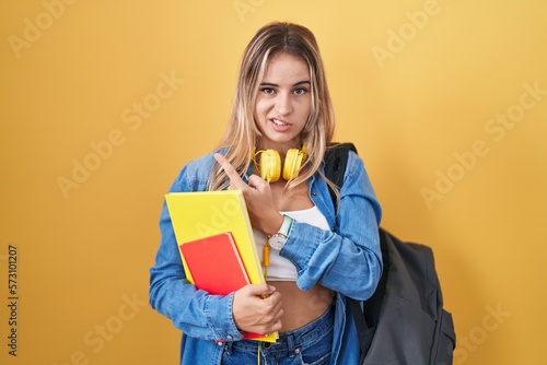 Young blonde woman wearing student backpack and holding books pointing aside worried and nervous with forefinger, concerned and surprised expression