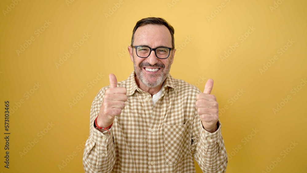 Middle age man smiling confident doing ok sign with thumbs up over isolated yellow background