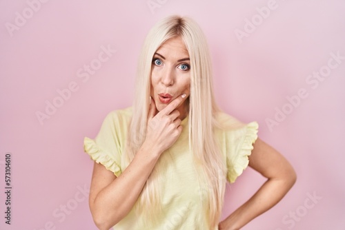 Caucasian woman standing over pink background looking fascinated with disbelief, surprise and amazed expression with hands on chin