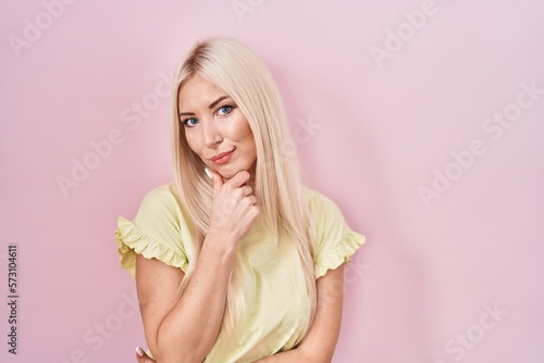 Caucasian woman standing over pink background looking confident at the camera smiling with crossed arms and hand raised on chin. thinking positive.