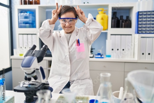 Hispanic girl with down syndrome working at scientist laboratory posing funny and crazy with fingers on head as bunny ears  smiling cheerful