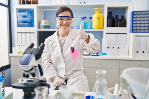 Hispanic girl with down syndrome working at scientist laboratory doing happy thumbs up gesture with hand. approving expression looking at the camera showing success.