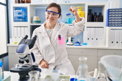 Fototapeta Naklejka Na Ścianę i Meble -  Hispanic girl with down syndrome working at scientist laboratory strong person showing arm muscle, confident and proud of power