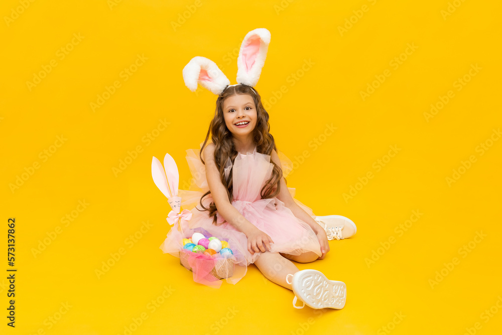 A little girl dressed as an Easter bunny with a basket of colorful eggs on a yellow isolated background. Festive spring Easter.