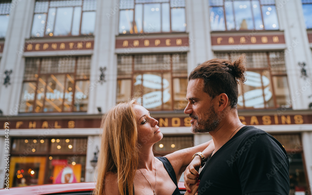 Young adult Hispanic beardy man embracing redhead girlfriend at street against blurry building. European couple dating. Relationship, romance. Handsome hipster guy with wife in city, travel.