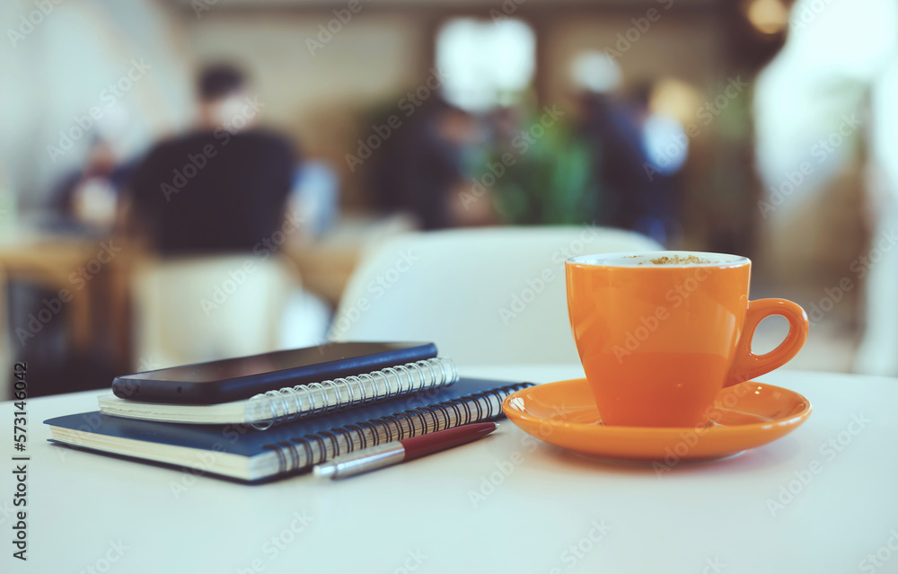Close-up view, oreange cup of coffee with smartphone, notebook and pen on white table in cafe, freelancer relax with coffee drink after work, blurred background, vintage collor style Close-up view, oreange cup of coffee with smartphone, notebook and pen on white table in cafe, freelancer relax with coffee drink after work, blurred background, vintage collor style