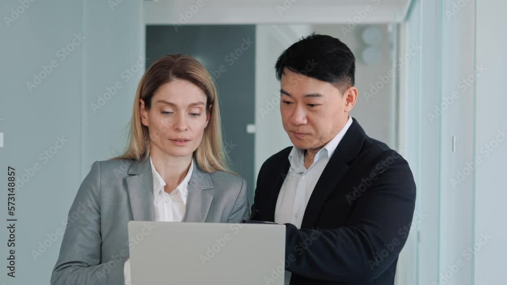 Multinational business partners working together looking at laptop screen. Two business people working in office hallway reading and discuss asian investment project, analyze the stock market. Multinational business partners working together looking at laptop screen. Two business people working in office hallway reading and discuss asian investment project, analyze the stock market.