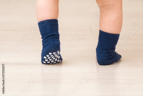 Baby boy legs on light beige wooden floor. Feet in dark blue anti slip socks. Infant first steps. Closeup. Back view. Safety walking on slippery floor at home. photo