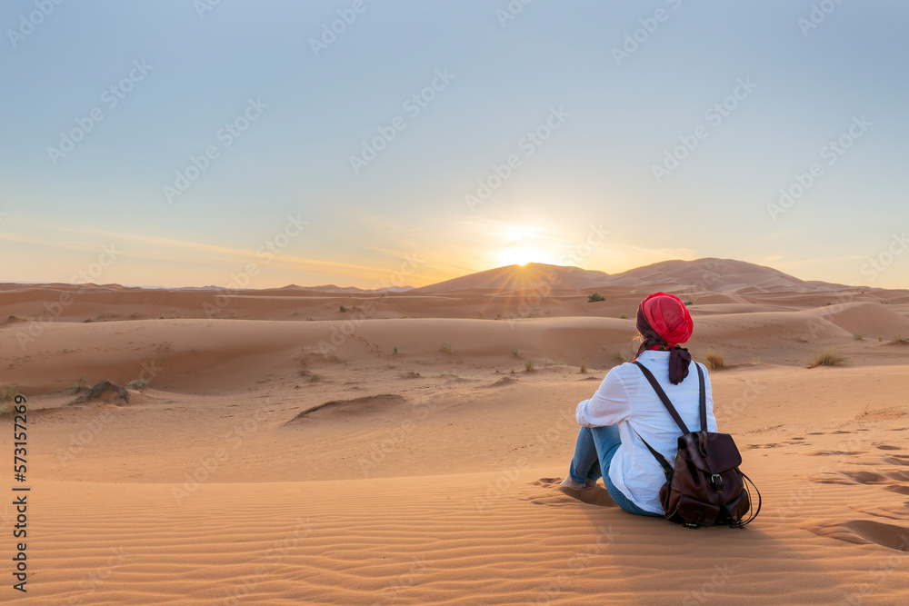 A young fermale traveler enjoys a sunrise or sunset landscape view of the desert sand dunes of Erg Chebbi near the village of Merzouga, Morocco. - obrazy, fototapety, plakaty 