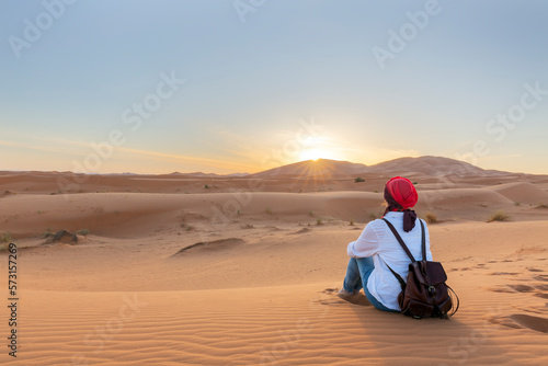 A young fermale traveler enjoys a sunrise or sunset landscape view of the desert sand dunes of Erg Chebbi near the village of Merzouga, Morocco.