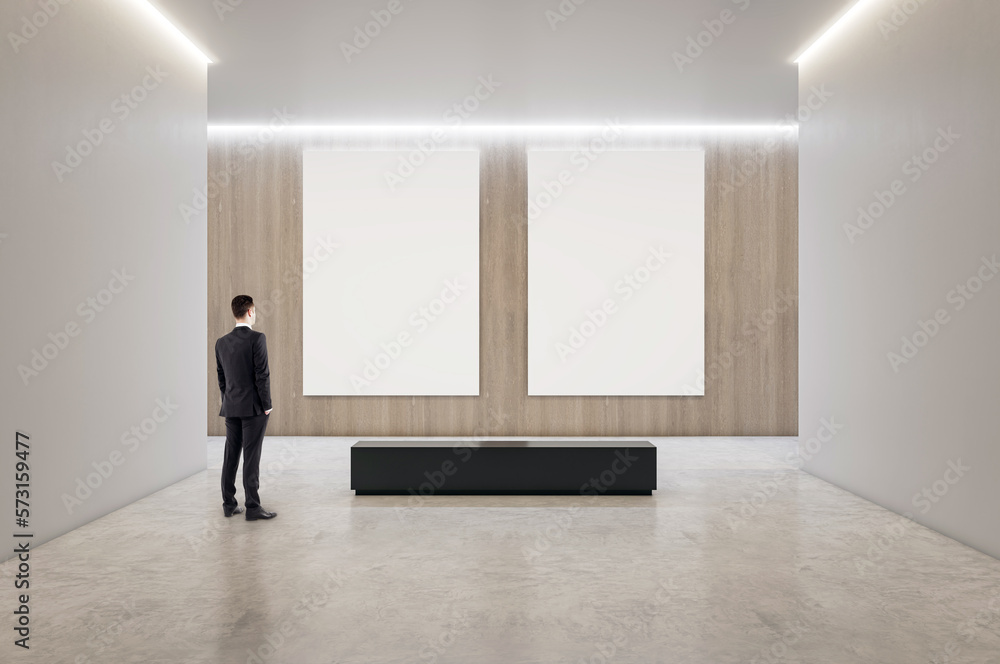 Back view on businessman looking at two blank white posters with space for your logo or text on light wooden wall background in spacious art center hall with black bench on concrete floor, mock up Back view on businessman looking at two blank white posters with space for your logo or text on light wooden wall background in spacious art center hall with black bench on concrete floor, mock up