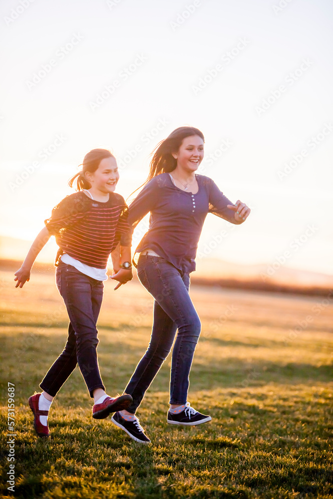 Two girls (sisters, one is a teenager and one is elementary aged) skip through a field at sunset while holding hands. Two girls (sisters, one is a teenager and one is elementary aged) skip through a field at sunset while holding hands.