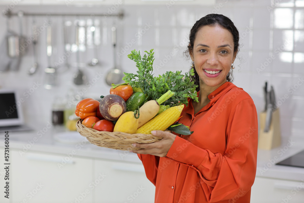 Happy latin woman holding a fruit basket for cooking while learning recipe from internet with mobile phone in kitchen. Young female preparing delicious meal at home. Healthy and lifestyle concept Happy latin woman holding a fruit basket for cooking while learning recipe from internet with mobile phone in kitchen. Young female preparing delicious meal at home. Healthy and lifestyle concept