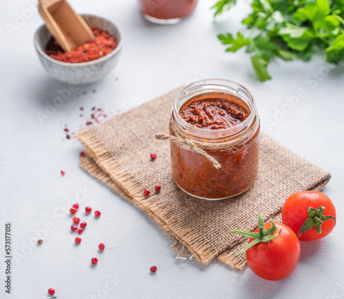 Hot sauce adjika. Homemade appetizer with pepper and tomatoes in a jar on light background with fresh herb and vegetables.