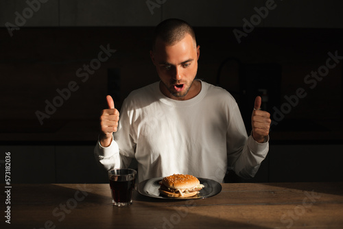 Attractive amazed surprised man show thumbs up gesture and sitting near table with plate with burger. Fast food. Male wear white long sleeves sitting at house in kitchen and look at burger, open mouth