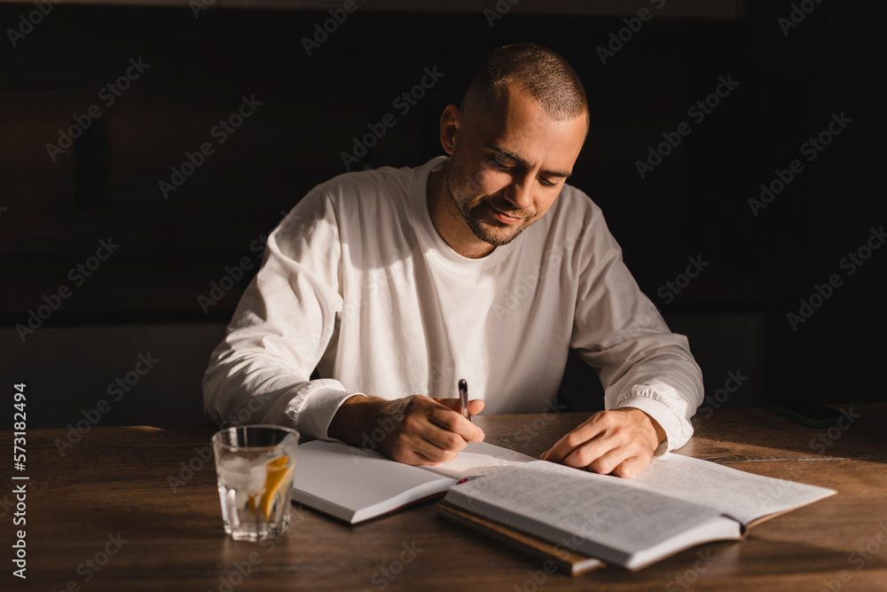 Man writing on paper notebook, look happy, smiling sitting at house. Man student writing in notebook on the table while studying at home. Male working distantly pay bills online prepare for test exam. Man writing on paper notebook, look happy, smiling sitting at house. Man student writing in notebook on the table while studying at home. Male working distantly pay bills online prepare for test exam.