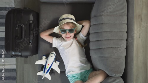 Smiling toothless little girl in sunglasses rests on sofa near airplane and suitcase at home. Happy preschooler ready for summer vacation upper closeup