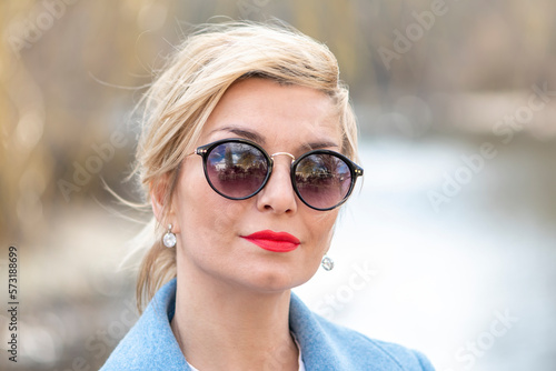 Street portrait of a stylish woman 40-45 years old in sunglasses on a blurry background of nature and water, close-up.