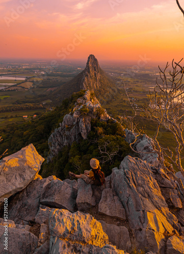 Picture from the back of a woman sitting on the peak of high mountain cliff. There are a lot of rock with white color. The sun is setting on the mountain and there is a beautiful warm orange light. © Pang wrp