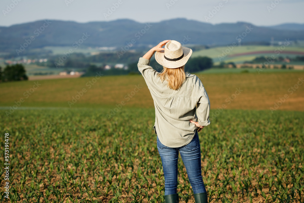 Woman with straw hat standing in agricultural field. Satisfied female farmer is looking at corn field in cultivated land Woman with straw hat standing in agricultural field. Satisfied female farmer is looking at corn field in cultivated land