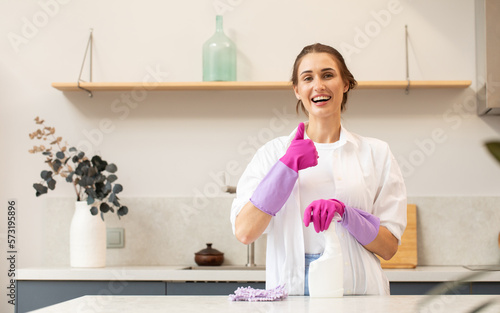 Attractive housewife holds a spray bottle with cleaning agent in her hand and shows a thumbs up. 