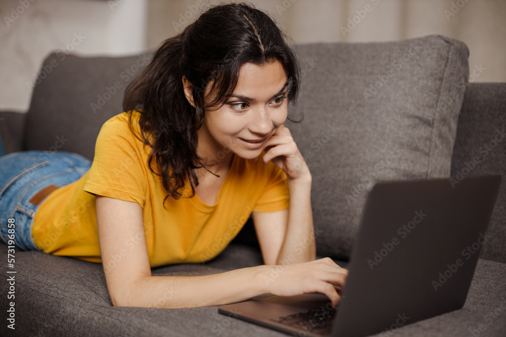 Fototapeta premium Happy indian or arabian young woman lying sofa and using laptop. Remote working