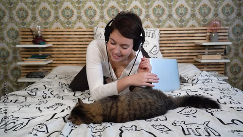 The beautiful woman is relaxing on the bed with her cat. The woman is lying down and she plays with the gray cat while she listens to music.
