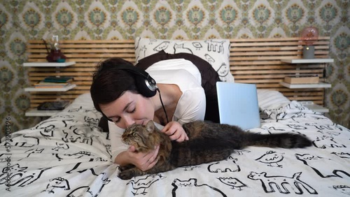 The beautiful woman is relaxing on the bed with her cat. The woman is lying down and she plays with the gray cat while she listens to music.

