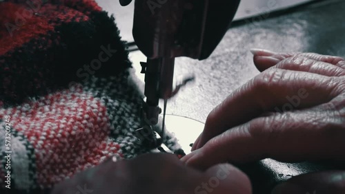 Aged woman sews on an old sewing machine. Needle and foot of sewing machine penetrates fabric with high magnification and super slow motion. Seamstress makes a machine-made even seam on red fabric.