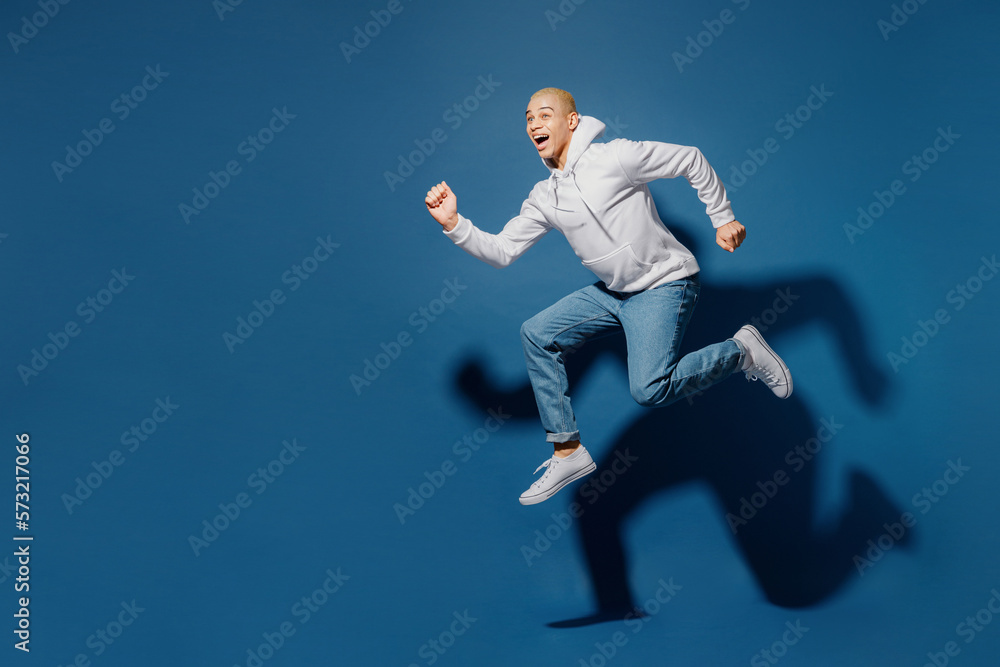 Fototapeta premium Full body side view sporty fun young dyed blond man of African American ethnicity in white hoody jump high run fast hurry up in rush isolated on plain dark royal navy blue background studio portrait