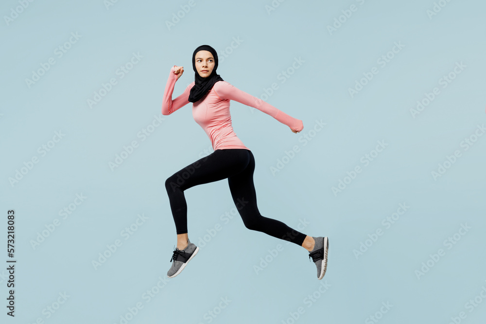 Full body sideways young asian muslim fitness trainer sporty woman wear pink abaya hijab spend time in gym run fast jump high isolated on plain blue background studio. Workout sport fit abs concept.