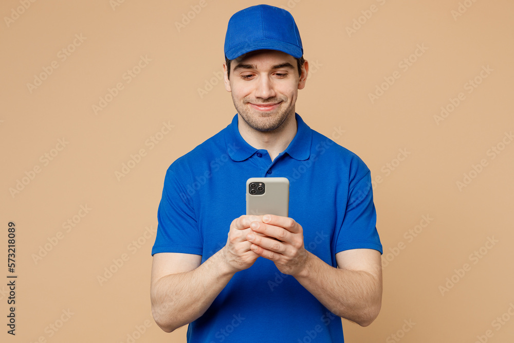 Professional fun delivery guy employee man wear blue cap t-shirt uniform workwear work as dealer courier hold in hand use mobile cell phone isolated on plain light beige background. Service concept. Professional fun delivery guy employee man wear blue cap t-shirt uniform workwear work as dealer courier hold in hand use mobile cell phone isolated on plain light beige background. Service concept.