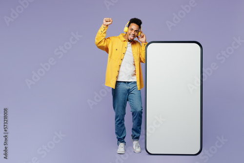 Full body young man of African American ethnicity wears yellow shirt t-shirt headphones big huge blank screen mobile cell phone smartphone with area listen music isolated on plain purple background.