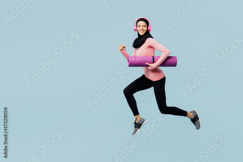 Fototapeta Naklejka Na Ścianę i Meble -  Full body young asian muslim fitness trainer sporty woman wear pink abaya hijab headphones hold yoga mat spend time in home gym isolated on plain blue background studio. Workout sport fit abs concept.