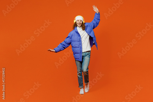 Young man with long curly hair wearing hat purple ski padded jacket casual clothes headphones listen to music dance raise up hands close eyes isolated on plain orange red background studio portrait.